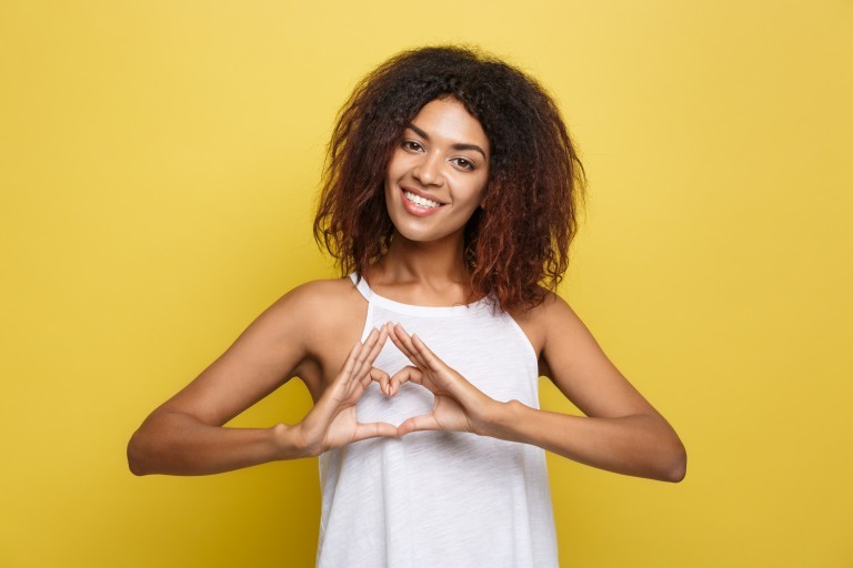 Isolated portrait of beautiful African American make heart symbol by hands. Yellow studio background. Copy Space.