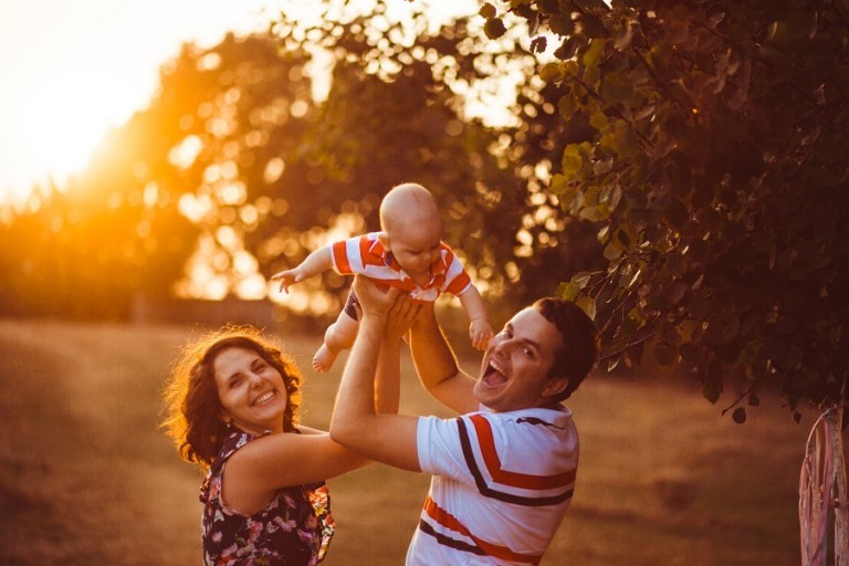 Cheerful parents hold their little son up standing outside in the evening