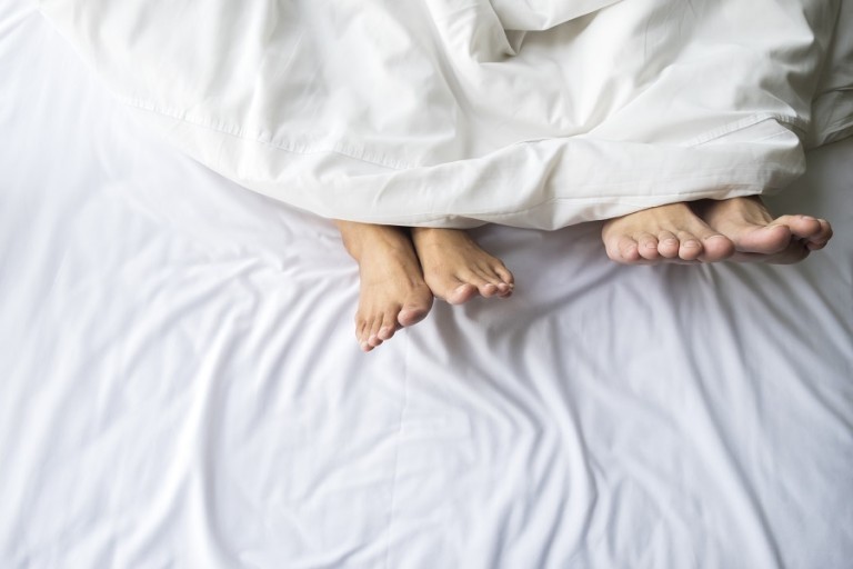 Feet of couple sleeping side by side in comfortable bed. Close up of feet in a bed under white blanket. Bare feet of a man and a woman peeking out from under the cover.Top view with background copy space.